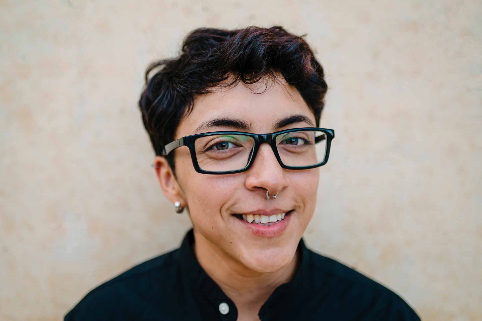 close up view of a young transgender man with black hair and glasses looking at the camera and smiling